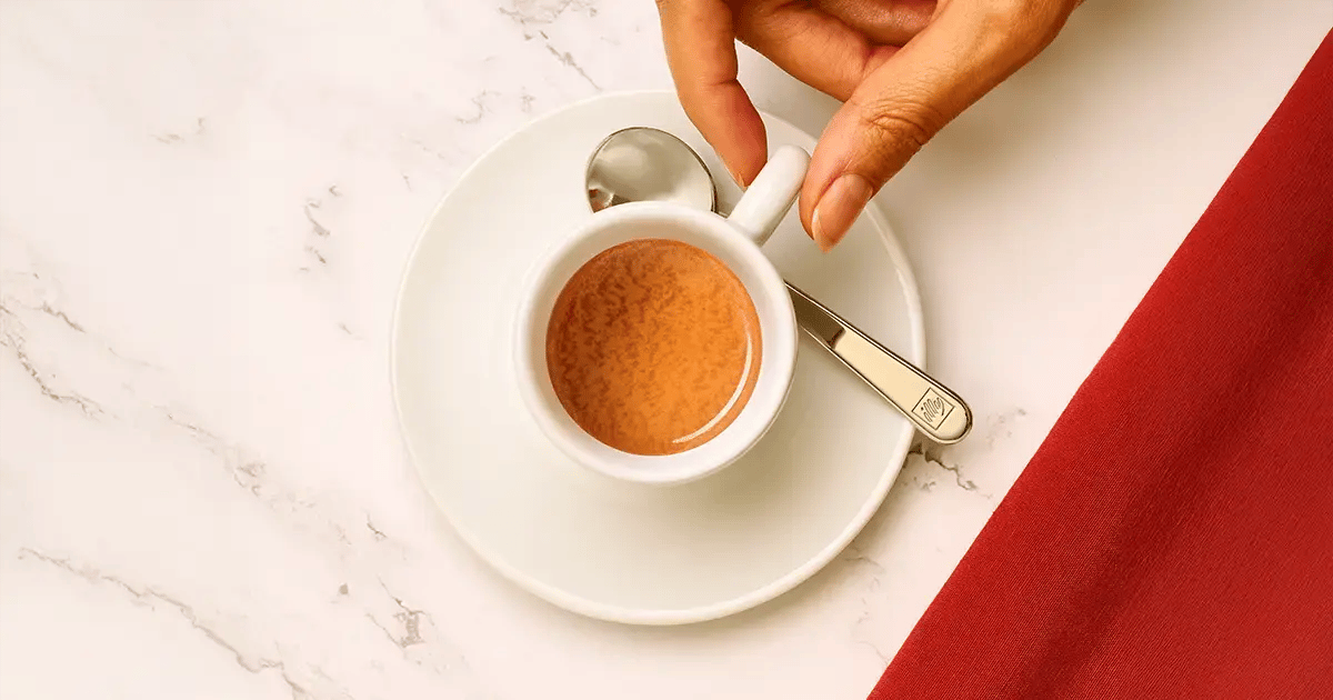 Mano che prende una tazzina di caffè illy dal piattino appoggiato su un tavolo illuminato da luce naturale.